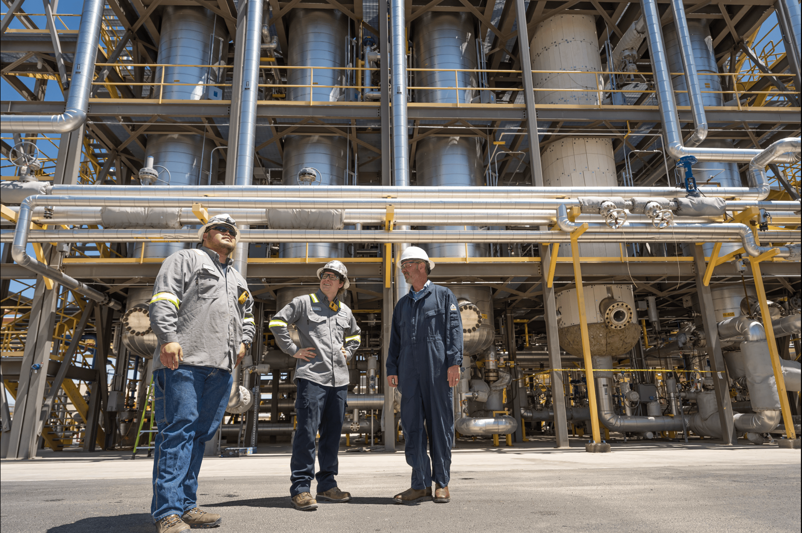 Three men stand in front of large refinery and distillery equipment