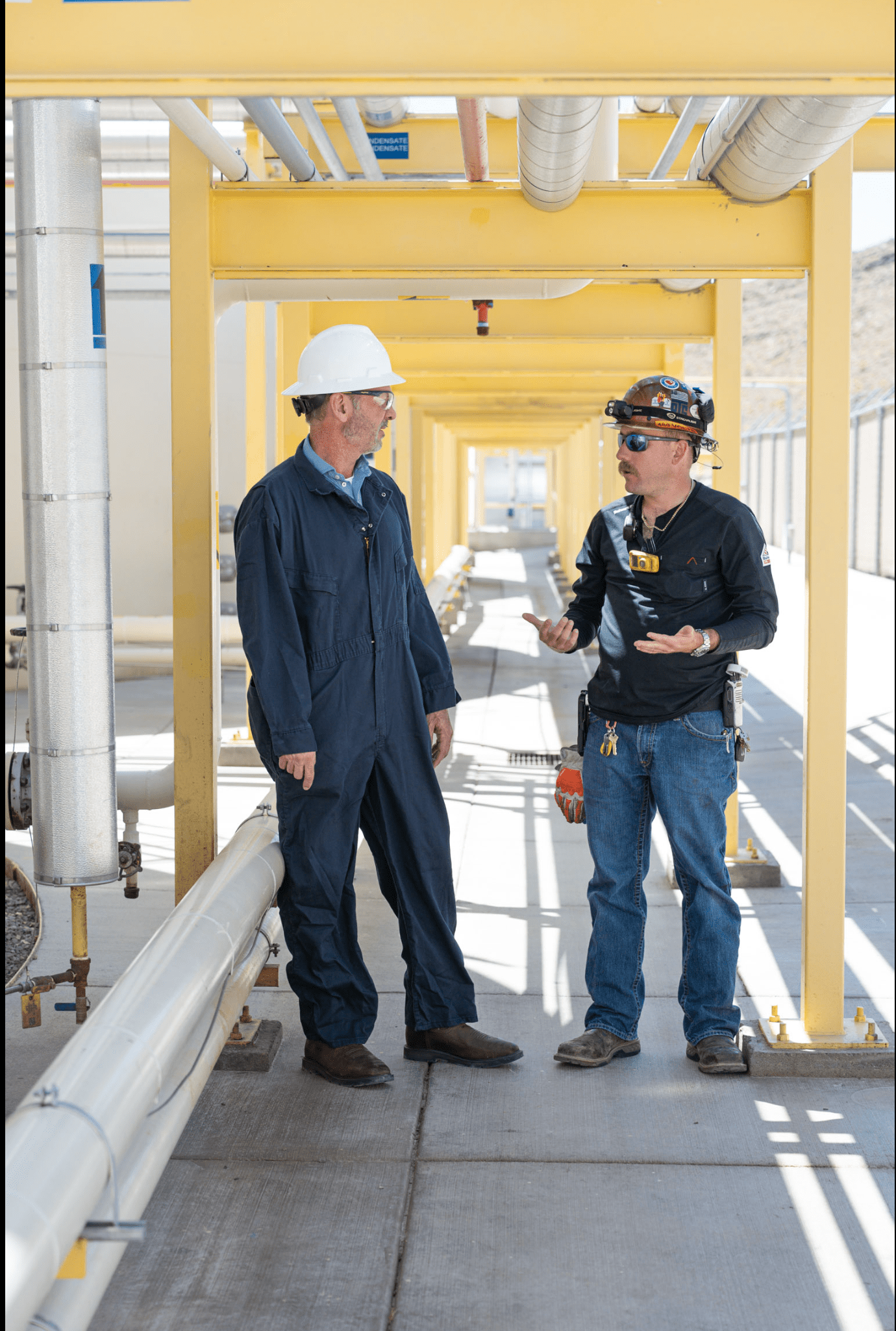 Two men wearing PPE stand under a fuel refinery