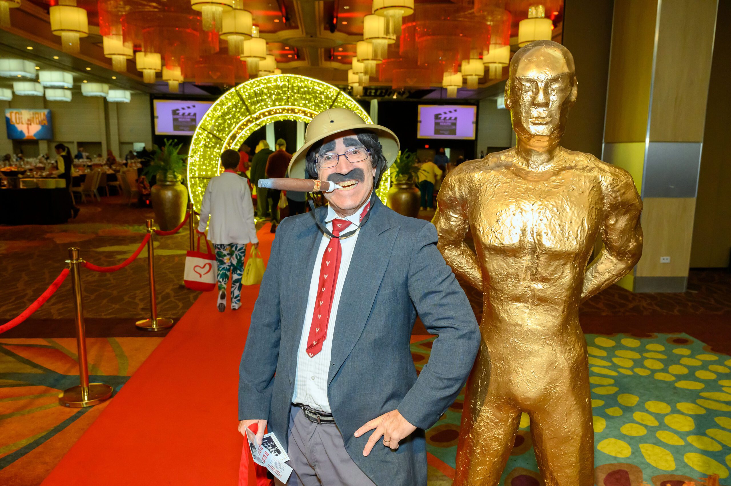 Man dressed as safari person with novelty cigar standing next to golden statue
